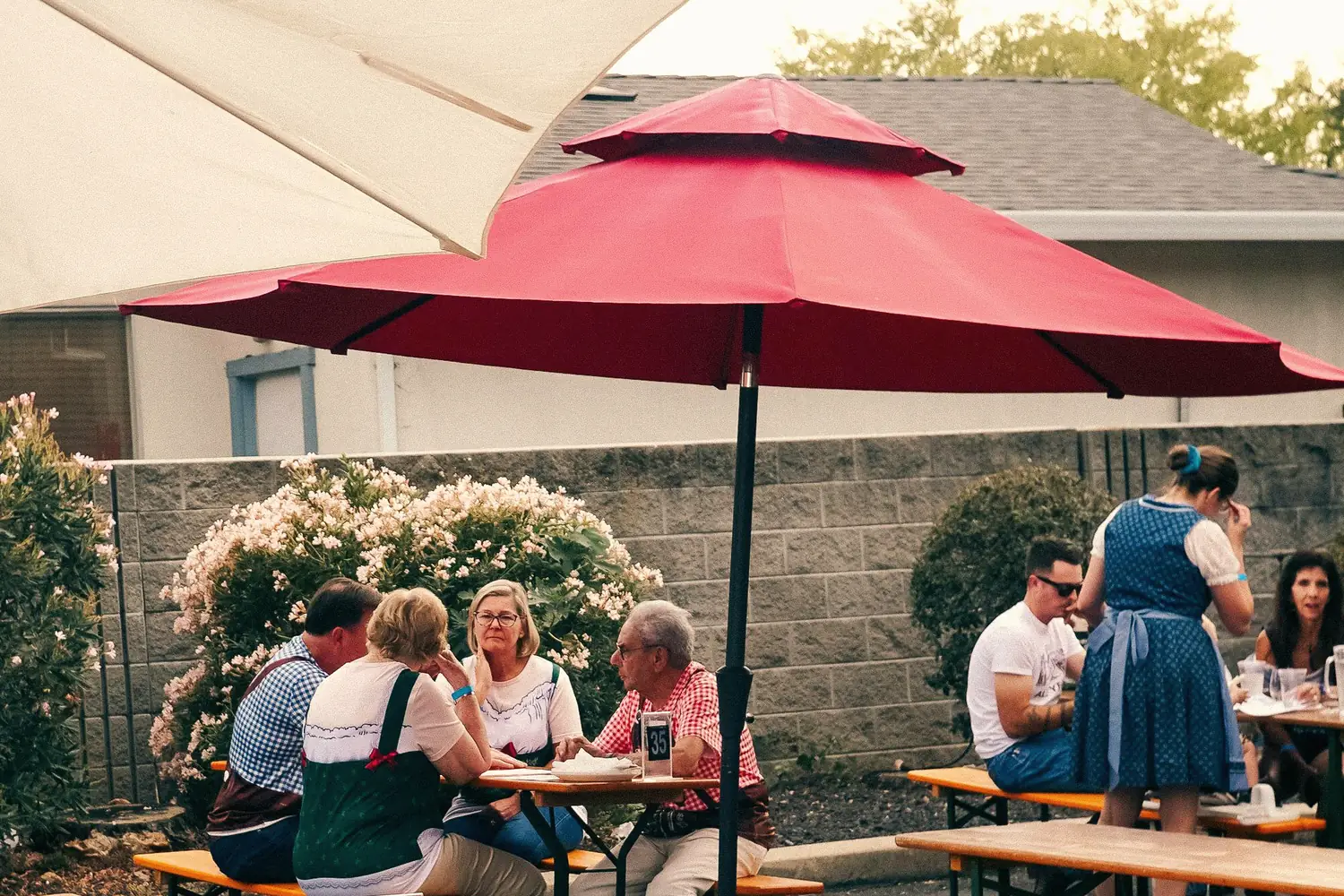 Endlose Biergarten-Tische unter grünen Bäumen, Sonnenschirme, Gäste.